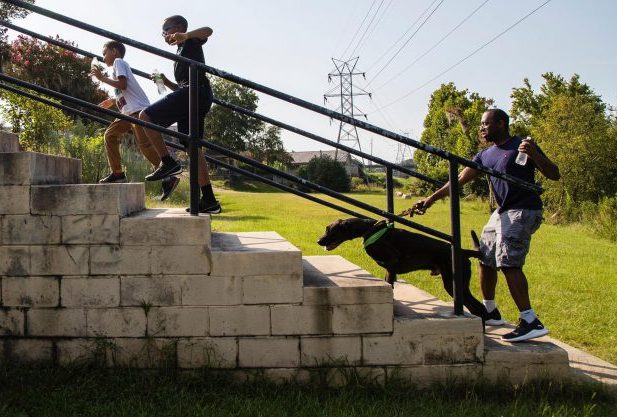 A Black family walks up the park stairs; two sons in front followed by their father with a dog on a leash and holding a water bottle