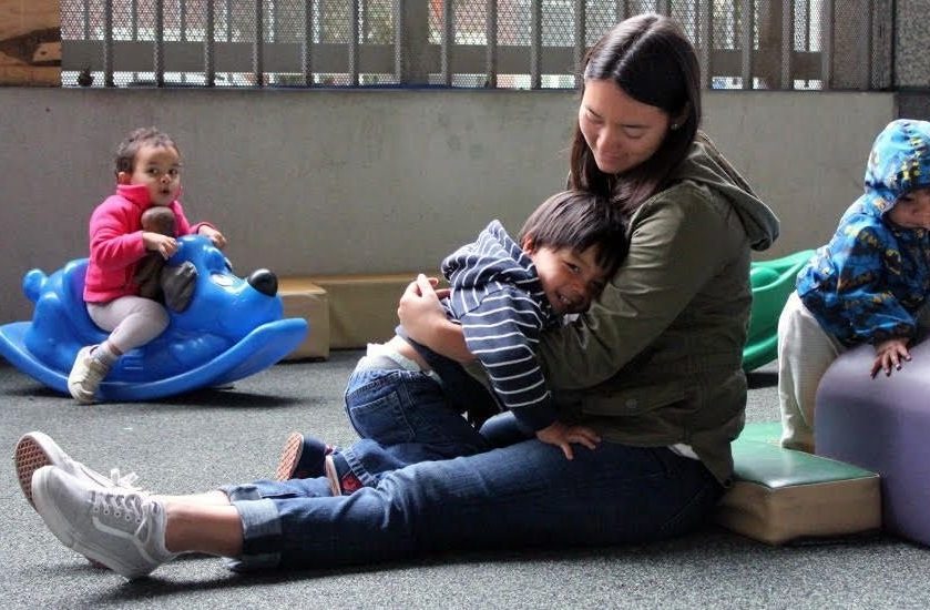 young mother with long dark hair sits on the floor with legs stretched out and little toddler boy snuggles on her lap