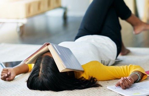 female college student lies back on bed surrounded by notebooks with open book resting on her face