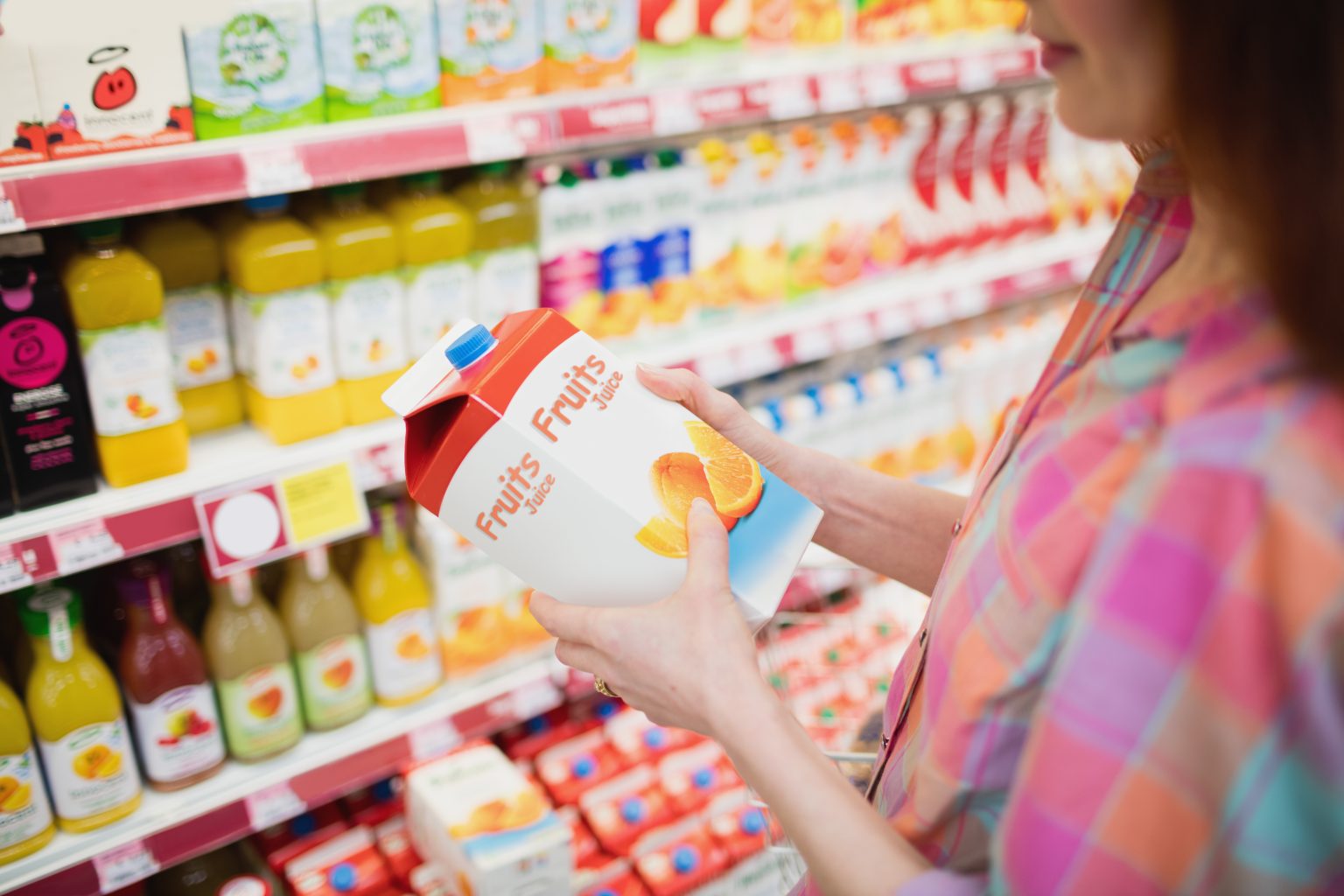 White woman with long brown hair wearing colorful plaid shirt stands in grocery store aisle holding carton reading 