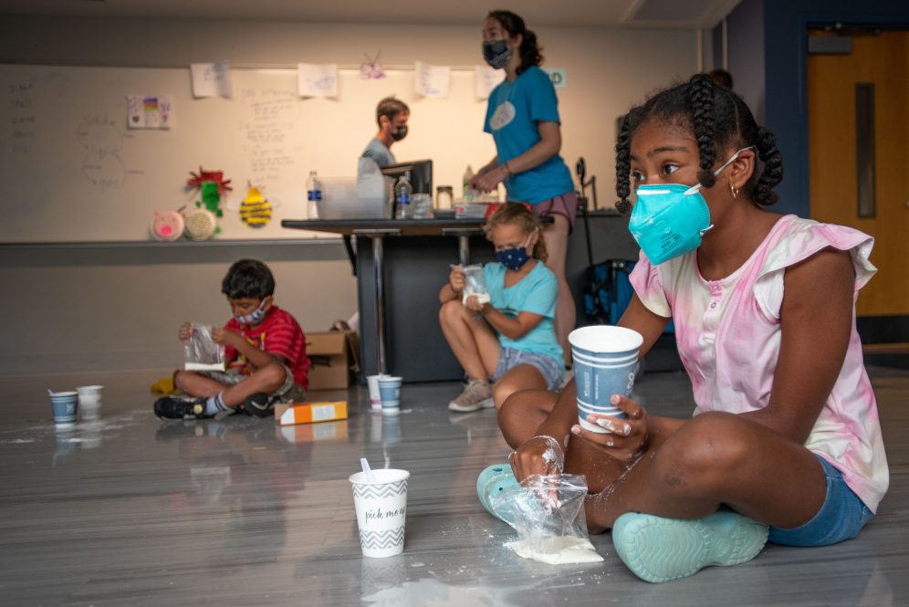Kids making oobleck at UConn BRAIN Camp; Black girl sits in foreground on floor adding baking soda to a cup