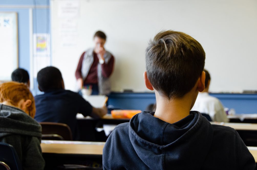 View from back of classroom, with teacher, standing with hand lifted to face, blurred in the background.