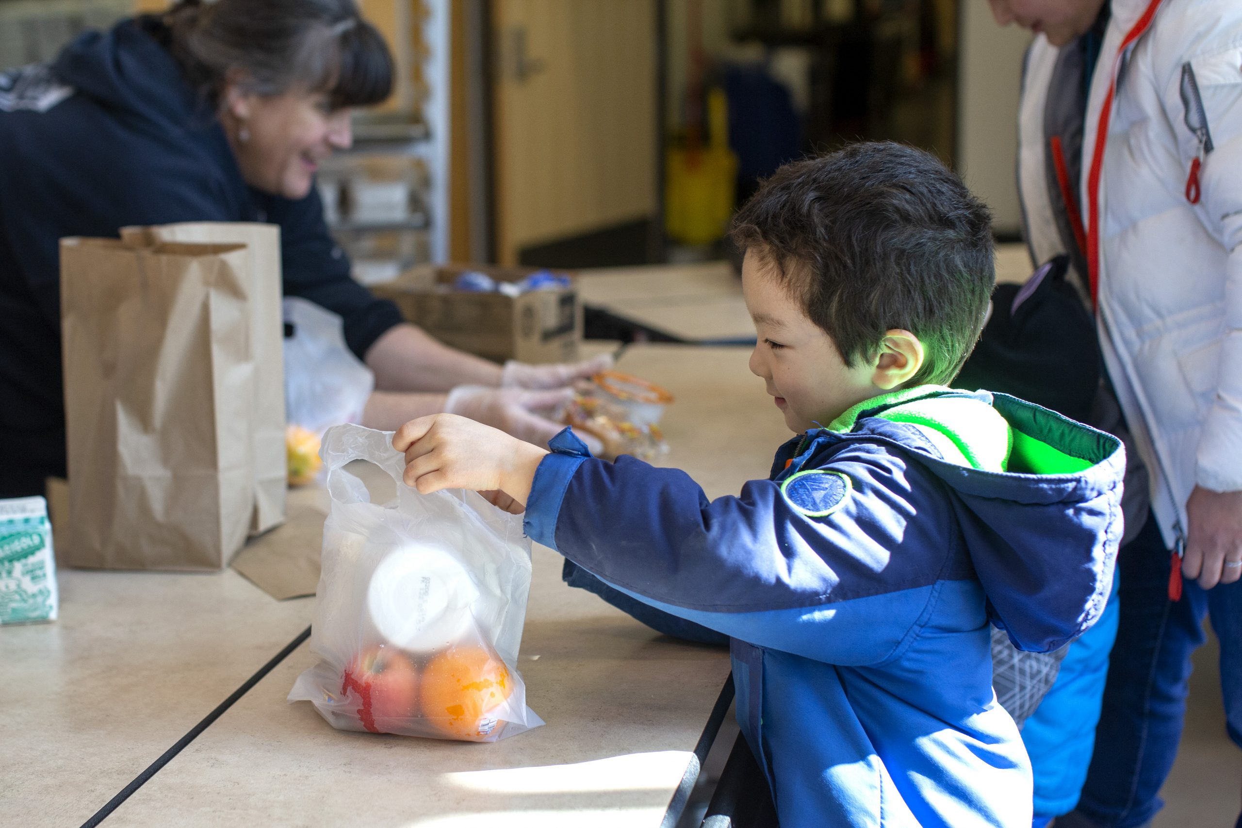 A young boy wearing a blue jacket picks up a bag of food from school