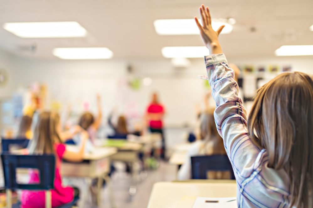 young child with long hair raises their arm in a school classroom. They are sitting at a desk. A teacher stands in front of students in a blurred background