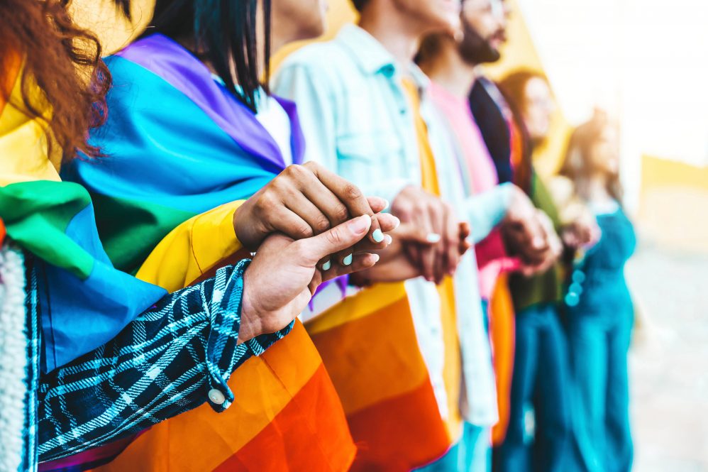 Line of people standing holding hands at pride event. The photo stops at chin level. One person is draped in rainbow flag.